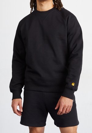 Mann trägt schwarzen Langarm-Sweatshirt mit gelbem Logo am Ärmel und schwarze Shorts vor weißem Hintergrund.