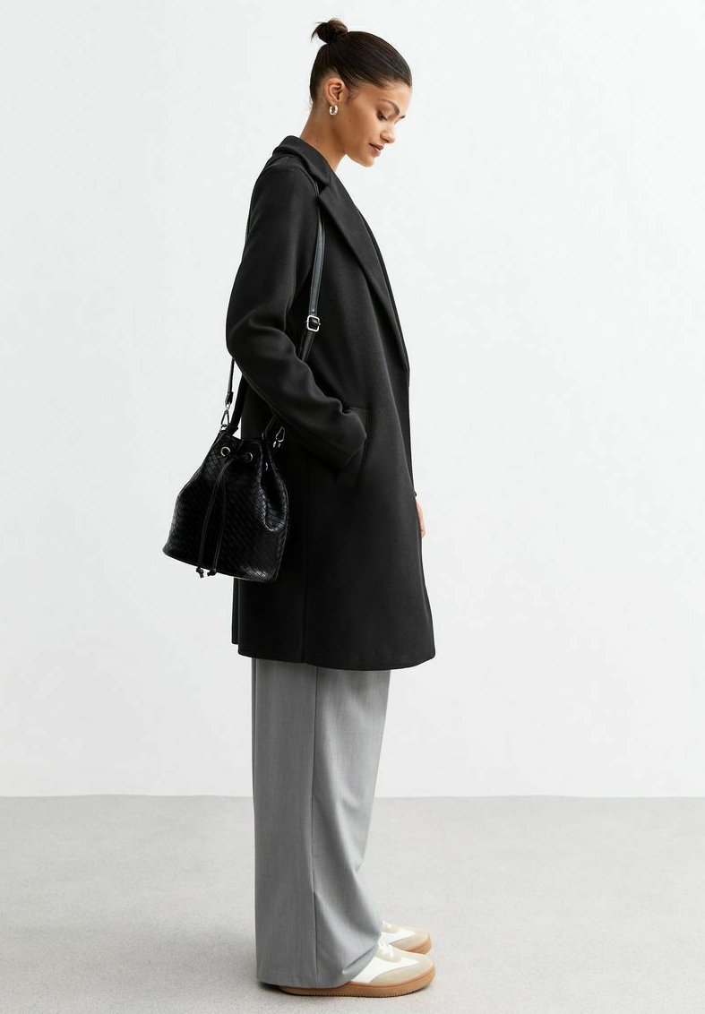 Un long manteau noir au design ajusté, un pantalon large gris clair et des chaussures à semelle beige. Un sac seau noir texturé avec cordon de serrage.