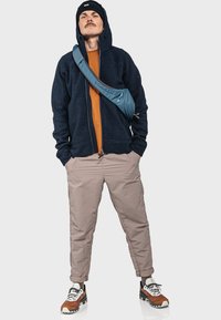 Navy Kapuzenpullover, orange Hemd, beige Hose, blaue Umhängetasche und braune/weiße Schuhe; lässiges Outfit mit lockerer Passform und unterschiedlichen Texturen.