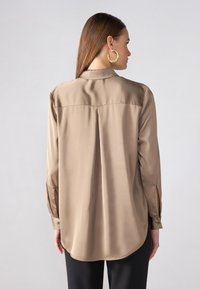 Beige Satinbluse mit langen Ärmeln, klassischem Kragen und einem gefältelten Rücken. Verfügt über geknöpfte Manschetten und seitliche Schlitze. Taillierte Passform.