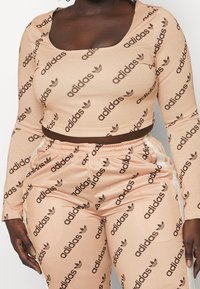 Detailní záběr osoby v broskvově zbarveném crop topu Adidas a odpovídajících kalhotách, obojí pokryté diagonálními černými logy Adidas a trifolovými symboly.