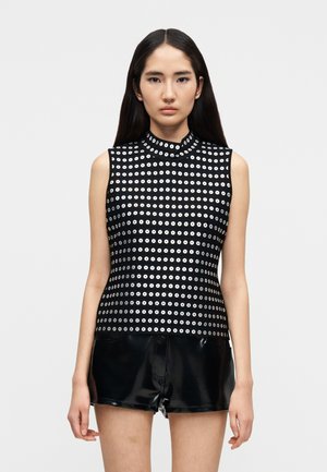 Mlada ženska z dolgimi temnimi lasmi, oblečena v črno brezrokavnik s srebrnimi krožnimi vzorci in sijoče črne kratke hlače, stoji pred belim ozadjem.