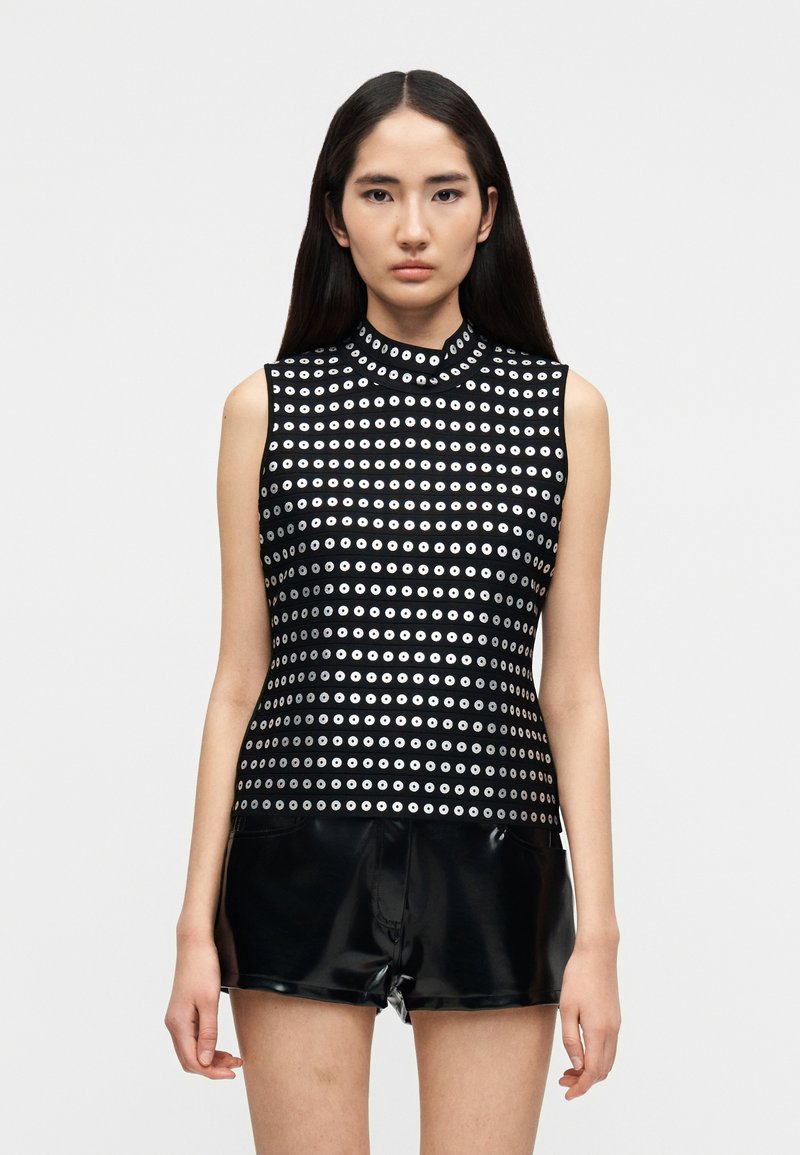 Νεαρή γυναίκα με μακριά σκούρα μαλλιά, φοράει αμ άνικο μαύρο τοπ με ασημένια κυκλικά σχέδια και λαμπερό μαύρο σορτς, στέκεται μπροστά σε λευκό φόντο.