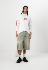 Hvid langærmet skjorte med rød grafisk print, parret med lyse denimshorts, sorte klumpede sko og holder en lærredstasker.