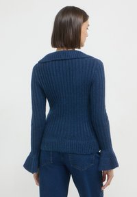 Femme aux cheveux courts bruns portant un pull côtelé bleu marine et un jean bleu foncé, de dos contre un fond clair uni.