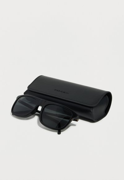 Μαύρα ορθογώνια γυαλιά ηλίου με σκουρόχρωμους φακούς τοποθετημένα μπροστά από μια μαύρη δερμάτινη θήκη με την επωνυμία Dolce & Gabbana, σε λευκό φόντο.