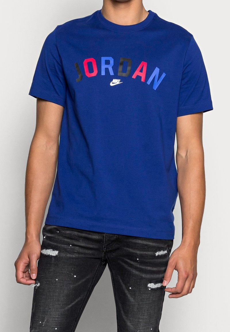 Hombre vestido con una camiseta azul de Jordan con letras multicolores y unos jeans negros desgastados con manchas de pintura blanca.