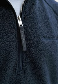 Prim-plan al unei jachete fleece bleumarin, cu fermoar argintiu și un șnur de prindere din material negru, inscripționat cu "Jack & Jones".