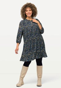Floral-patterned dress in navy blue with three-quarter sleeves, keyhole neckline, and flowing design. Worn with black leggings and beige boots.
