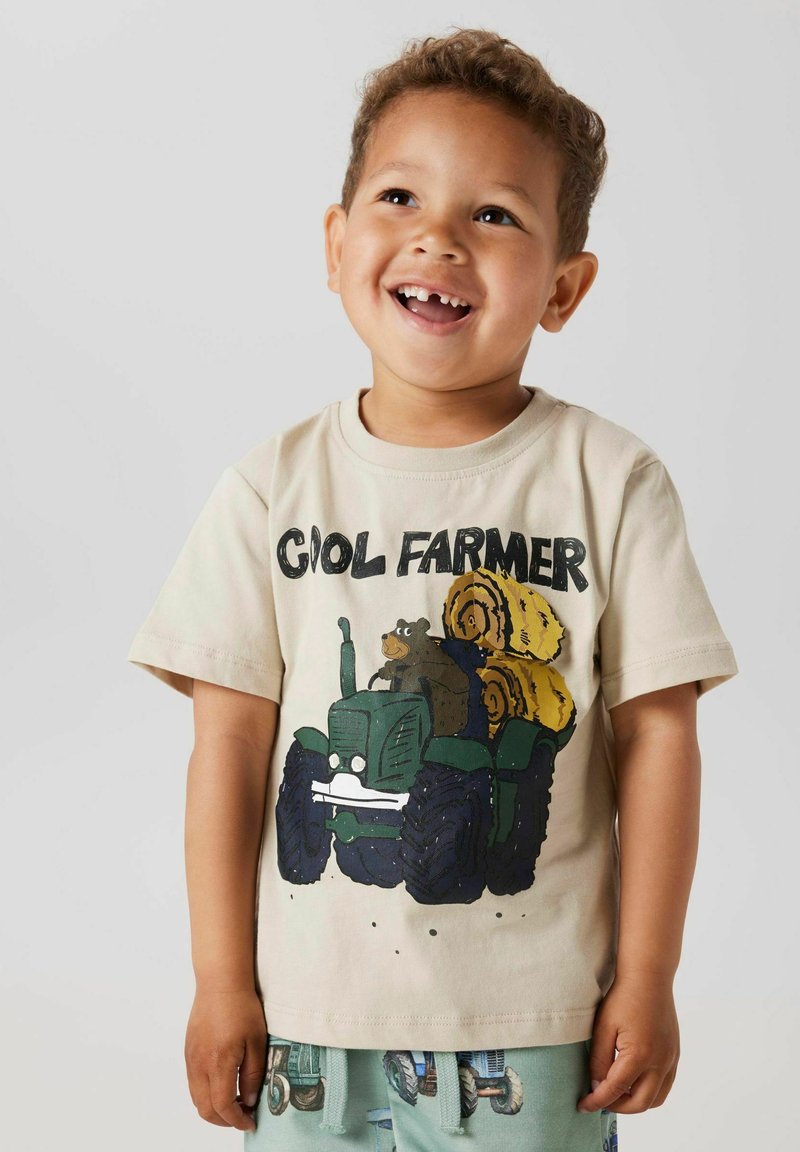 Camiseta de algodón color beige claro con un diseño de tractor y oso de dibujos animados en verde y amarillo. Mangas cortas, cuello redondo, corte relajado.