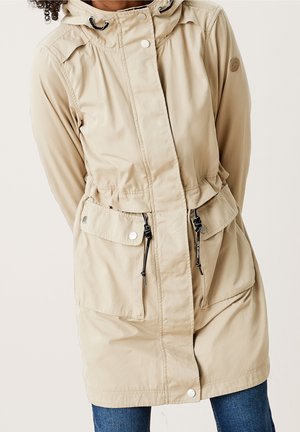 Veste beige à capuche avec boutons sur le devant, taille à cordon de serrage, poches à rabat, portée sur un jean bleu par une personne aux cheveux bouclés.