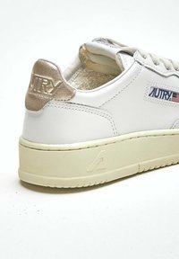Sneaker bianco in pelle con suola beige chiaro testurizzata, accentuazione del tallone in oro metallico e dettaglio del logo impresso sul lato.