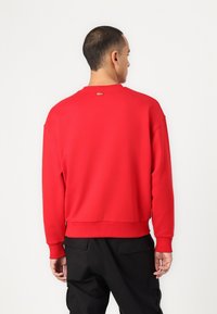 Felpa rossa con collo e orlo a costine. Accento del logo oro sul retro. Tessuto morbido con spalle scese. Pantaloni neri.