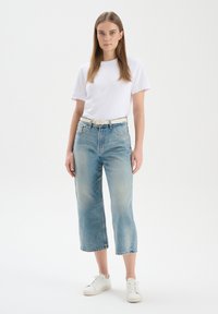 T-shirt bianca, jeans cropped azzurri chiari e sneakers bianche. I jeans hanno una vestibilità dritta e una leggera usura. La cintura è di colore chiaro.