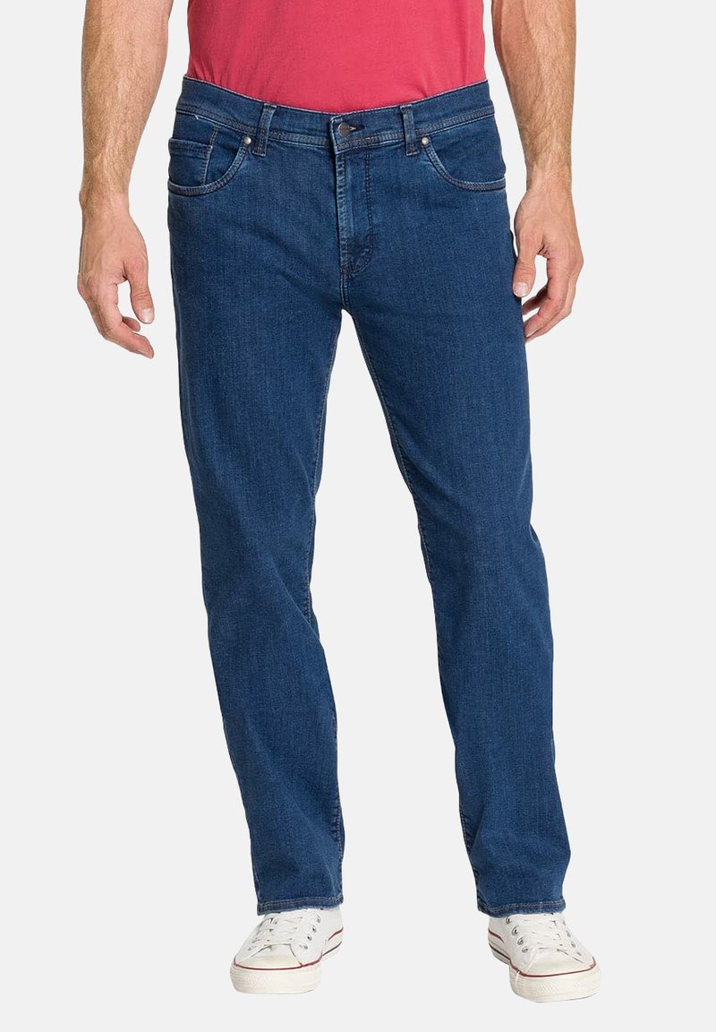 Homme portant un jean droit bleu, des baskets blanches et une chemise rouge, debout avec les bras relâchés le long du corps.