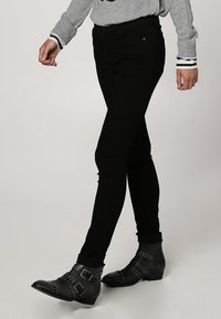 Jean noir avec une coupe slim, tissu robuste et légère brillance, assorti à des bottines noires avec boucles et clous.