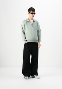 Joven con gafas de sol negras, sudadera de cuello con cremallera verde claro, pantalones anchos negros y zapatillas blancas de pie contra un fondo blanco.