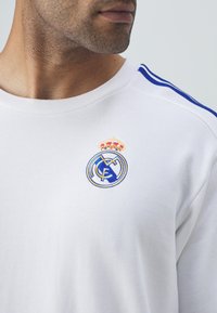 Fehér pamut pulóver, amelyen kék csíkok találhatók a vállakon, és hímzett Real Madrid logó látható egy koronával a mellen.