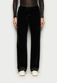 Černé sametové kalhoty s volným střihem, vybavené elastickým pasem a šňůrkou, kombinované s bílými teniskami. Hladký povrch a jednobarevný vzhled.