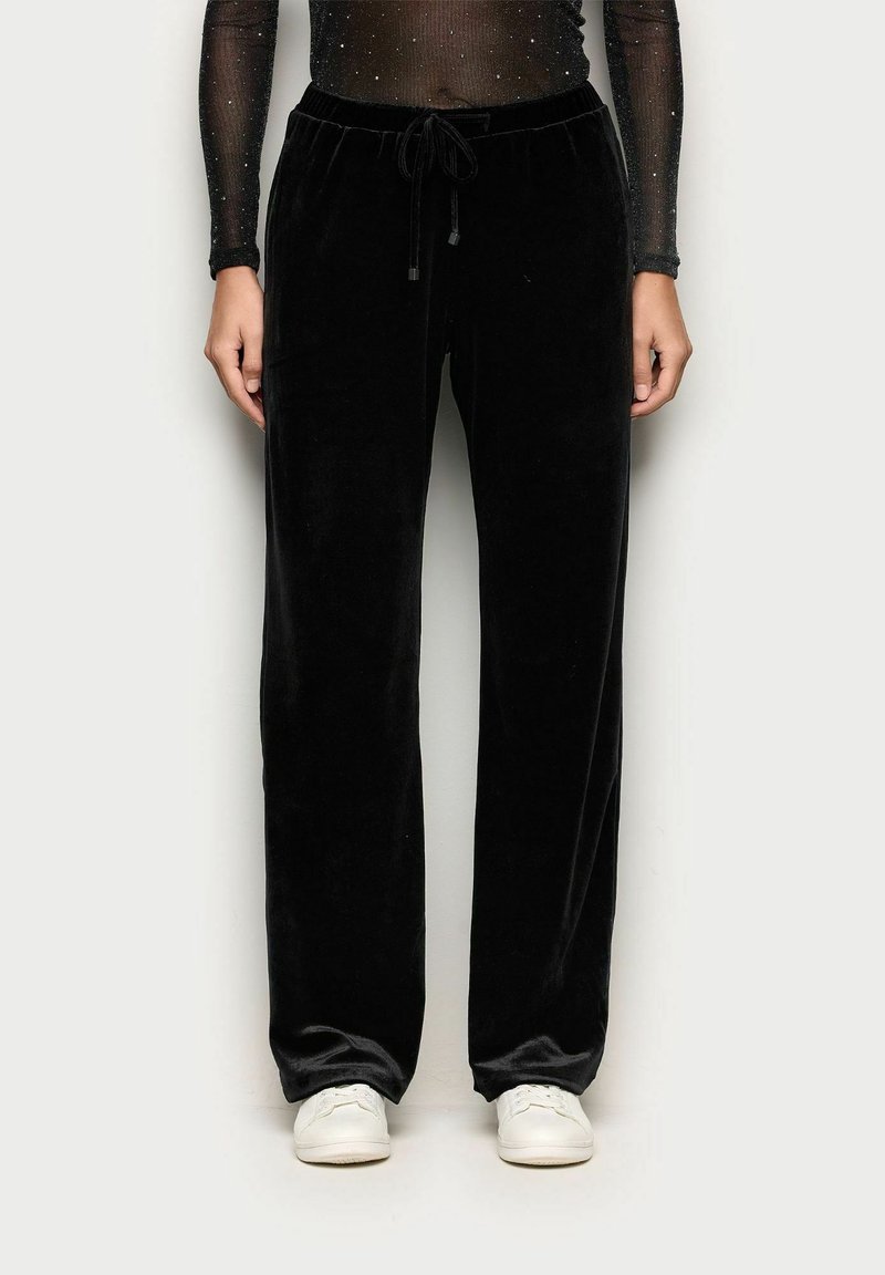 Černé sametové kalhoty s volným střihem, vybavené elastickým pasem a šňůrkou, kombinované s bílými teniskami. Hladký povrch a jednobarevný vzhled.