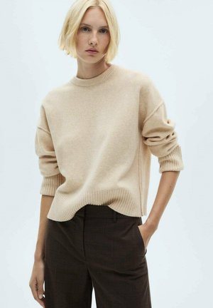 Pull en maille beige avec un col et des poignets côtelés, forme décontractée et épaules tombantes. Associé à un pantalon à carreaux marron.
