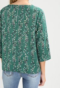 Grüne Bluse mit floralem Muster in Weiß und Lila, 3/4-Ärmel und Reißverschlussdetails auf dem Rücken. Getragen mit blauen Jeans.