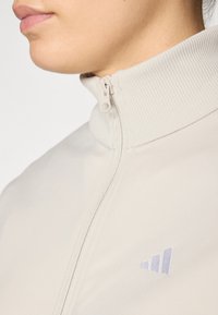 Lys beige zip-jakke med ribbet høykrage, med en liten lilla logo på brystet og et jevnt, teksturert stoff.