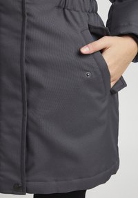 Vêtements d'extérieur texturé gris avec une taille élastique, des poches latérales et un détail de bouton-pression à l'ourlet. Présente une finition en tissu lisse.