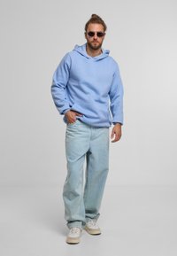 Felpa azzurra con cappuccio, tasca frontale, abbinata a jeans azzurri larghi e sneakers beige. Gli accessori includono occhiali da sole rotondi.
