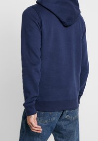 Man med en enfärgad marinblå hoodie och blå denimjeans, sedd bakifrån med synliga handtatueringar på vänster hand.