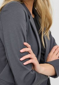 Szary, dopasowany żakiet o gładkiej fakturze tkaniny i usztywnionych ramionach. Posiada wąskie rękawy i subtelny połysk. Ręce skrzyżowane z przodu.