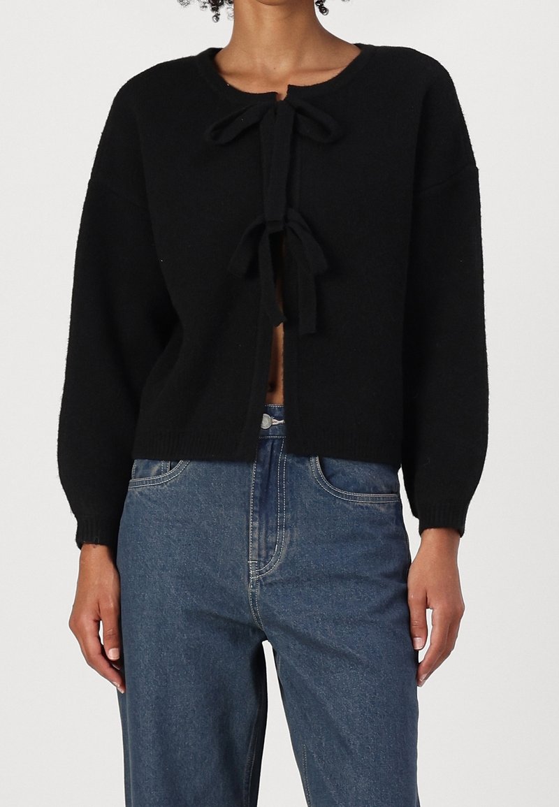 Cárdigan de punto negro con cuello redondo, mangas largas y lazos frontales. Combinado con jeans de mezclilla, que muestran un ajuste relajado.