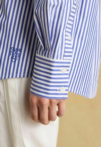 Ruka odpočíva na boku, oblečená v modro-bielom vertikálne pruhovanom dlhom rukáve s gombíkovými manžetami a bielych nohaviciach.