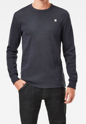 Uomo che indossa una maglia a maniche lunghe a coste di colore grigio scuro con un piccolo logo bianco G-Star RAW sul petto sinistro e pantaloni neri.