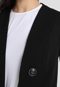 Černé sako s texturovaným povrchem, s jedním kulatým zapínac�ím knoflíkem a elegantním otevřeným střihem přes bílé tričko.