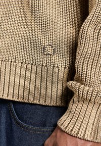 Beige knitted sweater featuring a ribbed hem and cuffs, with a small metal logo accent. Smooth texture with a warm, casual design.
