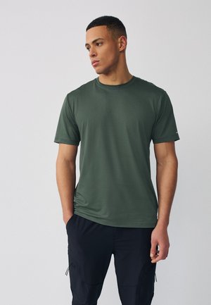 Muž v tmavozelenej tričku s krátkym rukávom a čiernych cargo nohaviciach, stojaci s rukami v kapsách, pozerajúci sa naľavo na jednoduchom pozadí.