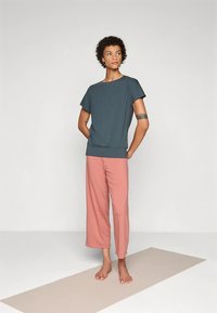 Person standing barefoot on a beige mat, wearing a dark teal short-sleeve top and loose pink pants, looking slightly to the side.