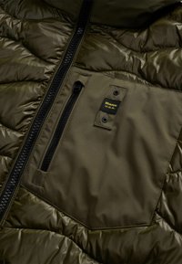 Olijfgroene gewatteerde jas met een glanzende afwerking, voorzien van een ritszak, zwarte rits en een merketiket op het borstgedeelte.