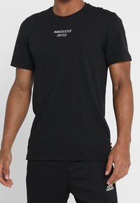 Czarna bawełniana koszulka T-shirt z krótkim rękawem, okrągłym dekoltem i białym nadrukiem "Manchester United" z przodu. Posiada regularny krój.