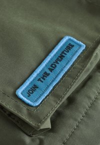 Parche de tela azul con texto negro "JOIN THE ADVENTURE" cosido sobre tela verde con costuras visibles.