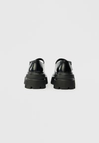 Sapatos de couro preto com sola preta robusta, apresentando uma textura suave e design arredondado. As traseiras possuem um pequeno detalhe em forma de alça para facilitar o calçado.