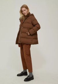 Giacca puffer marrone con cappuccio, dotata di chiusura con zip e bottoni a pressione. Il modello indossa pantaloni in velluto a coste marroni e mocassini neri con calzini grigi.
