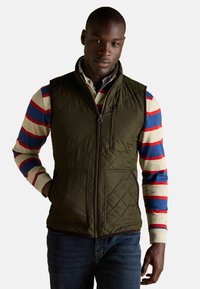 Olivgrüner, gesteppteter Weste mit hohem Kragen, Reißverschlusstaschen und einem Frontreißverschluss. Getragen über einem mehrfarbigen, gestreiften Langarmshirt.