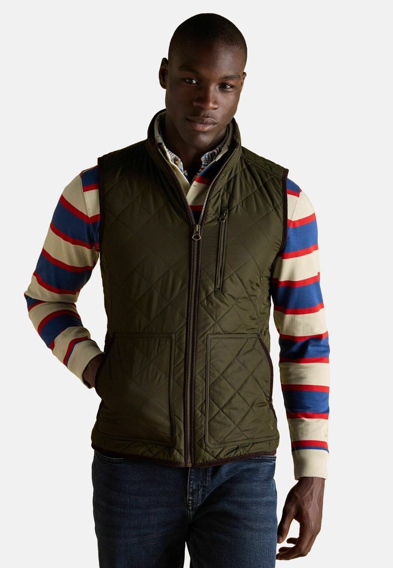 Olivgrüner, gesteppteter Weste mit hohem Kragen, Reißverschlusstaschen und einem Frontreißverschluss. Getragen über einem mehrfarbigen, gestreiften Langarmshirt.