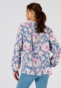 Blusa floral de manga larga en azul claro con flores rosas y blancas, con escote fruncido y detalles de puños elásticos. Llevada con jeans azules.
