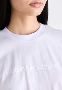 Bílý bavlněný tričko s klasickým kulatým výstřihem, s jemným bílým textem "Calvin Klein Jeans" napříč přední částí.
