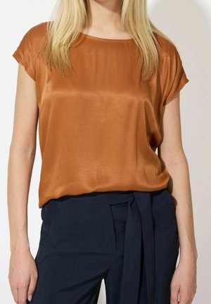 Kvinde iført en kortærmet kobberfarvet satinbluse stoppet ned i højtaljede marineblå bukser med stofbælte, stående mod en hvid baggrund.