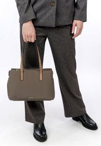 Persona con traje gris sostiene un bolso topo con asas color beige, usando botas de cuero negras sobre un fondo blanco.