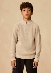 Beiger, gerippter Strickpullover mit einem halben Reißverschluss am Kragen und Raglanärmeln. Verfügt über eine strukturierte Oberfläche und einen umgeschlagenen Saum. Getragen mit dunklen Cargo-Hosen.